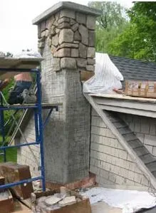 Chimney repair: Mason applying stones to chimney on a rooftop next to white shingle siding; construction scaffold visible.