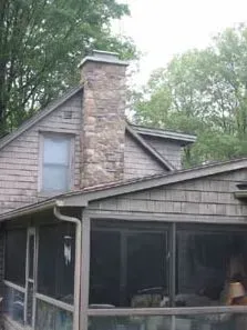 Stone chimney on a two-story house with a screened porch and gray shingle siding, surrounded by trees.