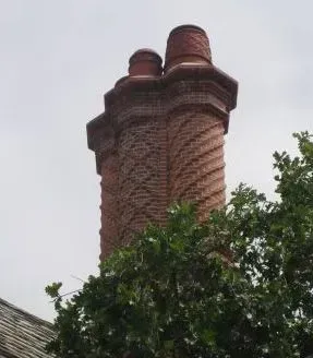 Tall, ornate brick chimney with two pots, swirling pattern, against a cloudy sky, partially obscured by green leaves.