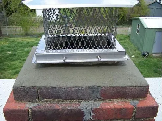 Chimney with metal cap and wire mesh on a brick structure, grass background.