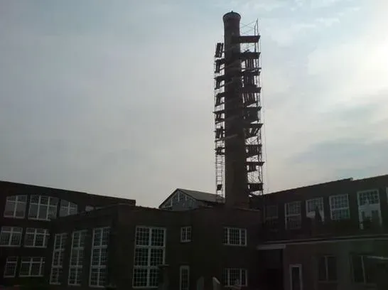 Brick factory building with tall chimney, scaffolding on chimney, cloudy sky.