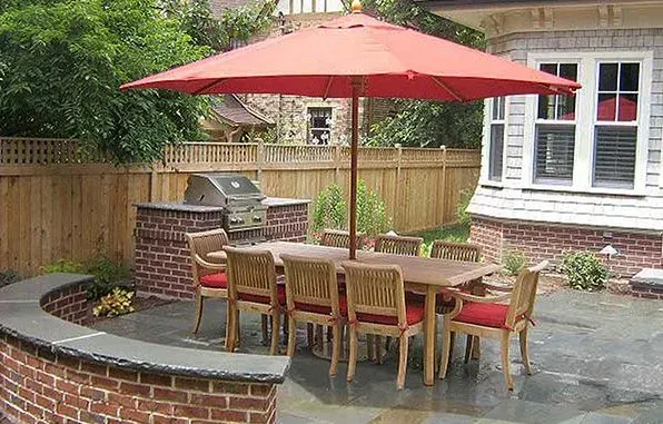 Outdoor dining area with red umbrella, wooden table and chairs, brick grill, and stone patio.