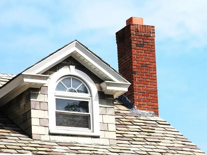 Dormer window with arched top and brick chimney against a blue sky.