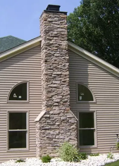 Stone chimney on a light-colored house with arched and rectangular windows, set against a blue sky.
