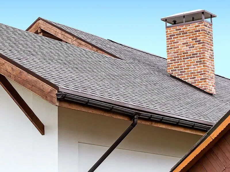 Close-up of a house roof with asphalt shingles, brown trim, brick chimney, and black gutters against a blue sky.
