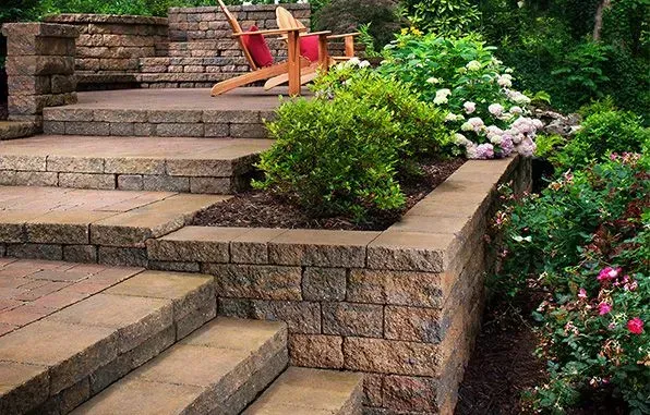 Stone patio with steps, retaining wall, and lush landscaping, including chairs on the upper level.