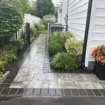 A paved walkway leads to a house; brick borders frame the path, greenery surrounds it.