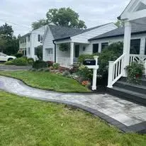 A house with a white fence and a mailbox on a paved path, surrounded by grass and plants.