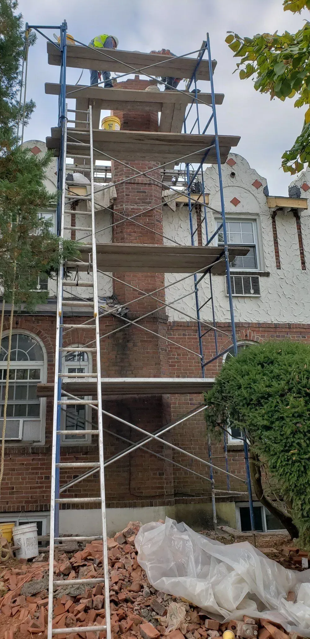 Brick chimney being repaired, scaffolding in place. Worker on top. Brick debris on ground.