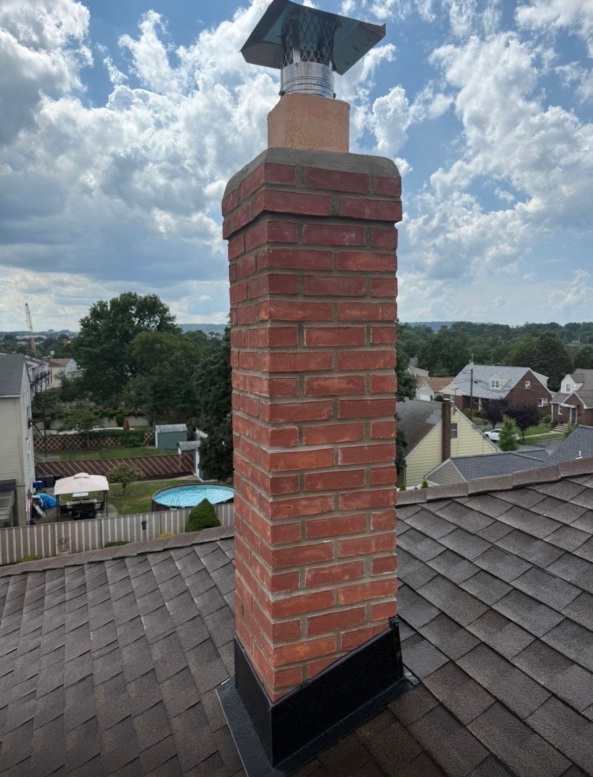Brick chimney on a shingled roof with a chimney cap, blue sky and residential neighborhood in background.
