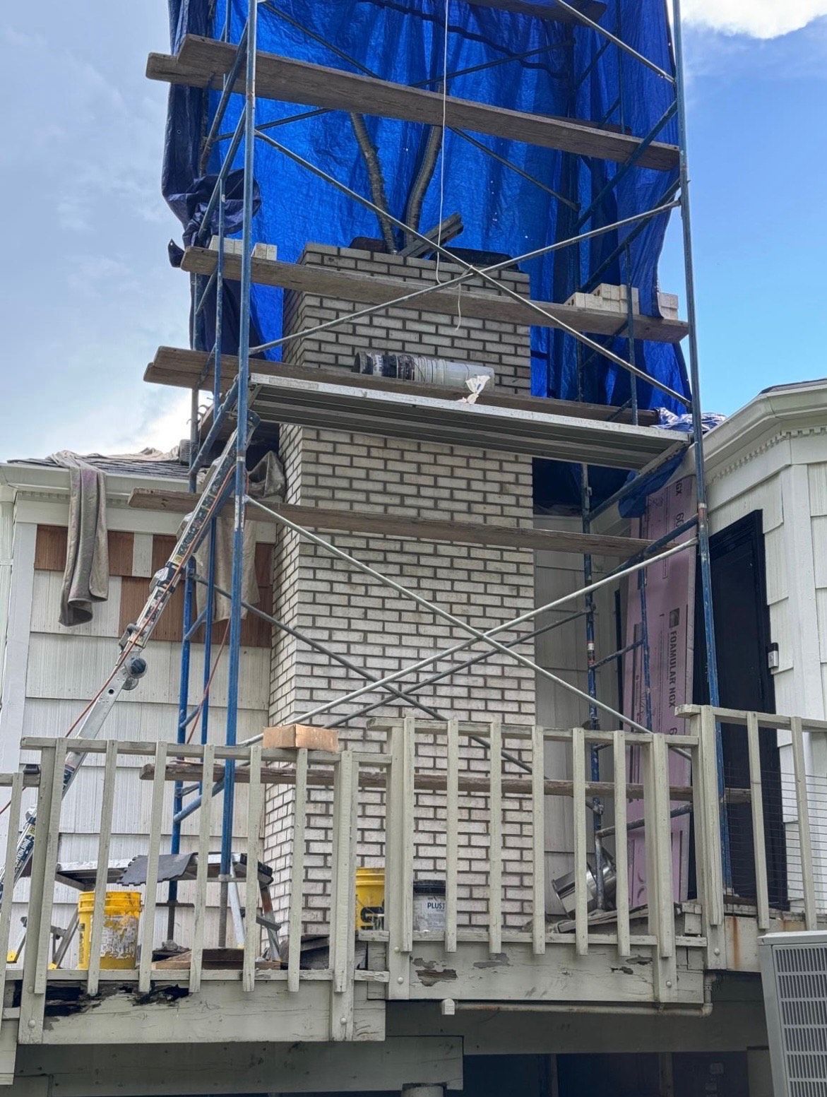 Chimney under construction with scaffolding and blue tarp, on a deck of a white house.