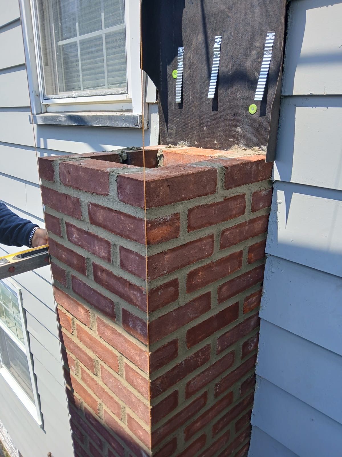 Brick chimney being built against a blue-sided house, black flashing installed.