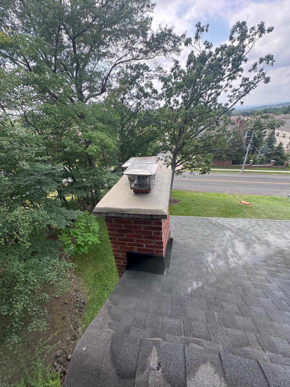 Brick chimney with a metal cap on a gray shingled roof, trees and street in the background.