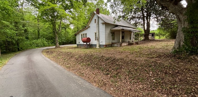 A small white house is sitting on the side of a road surrounded by trees.