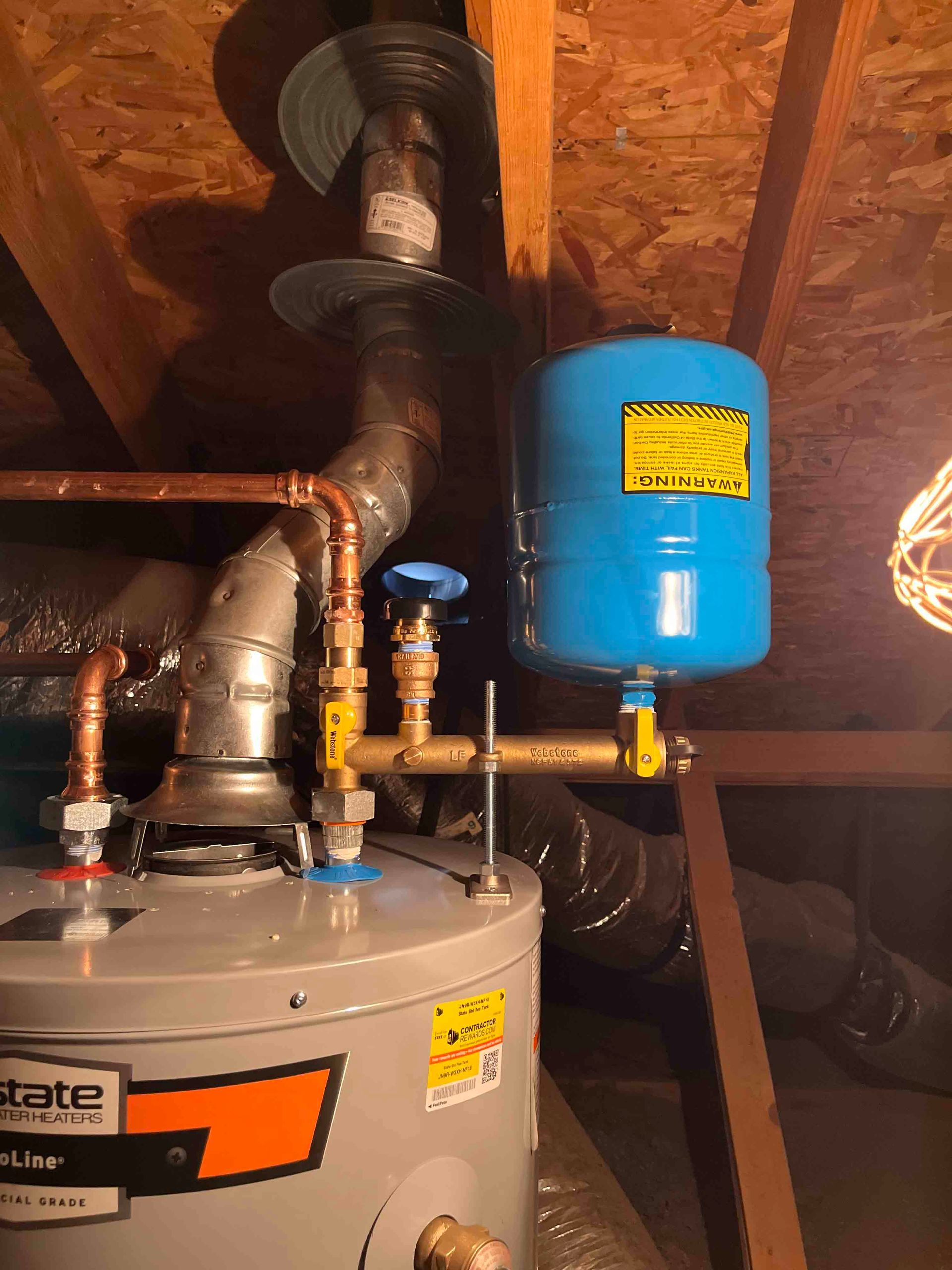 A water heater is sitting in an attic next to a blue tank.