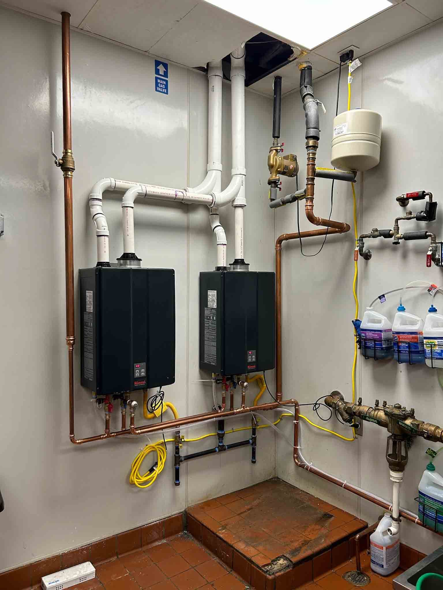 A room with two water heaters and a lot of pipes hanging from the ceiling.