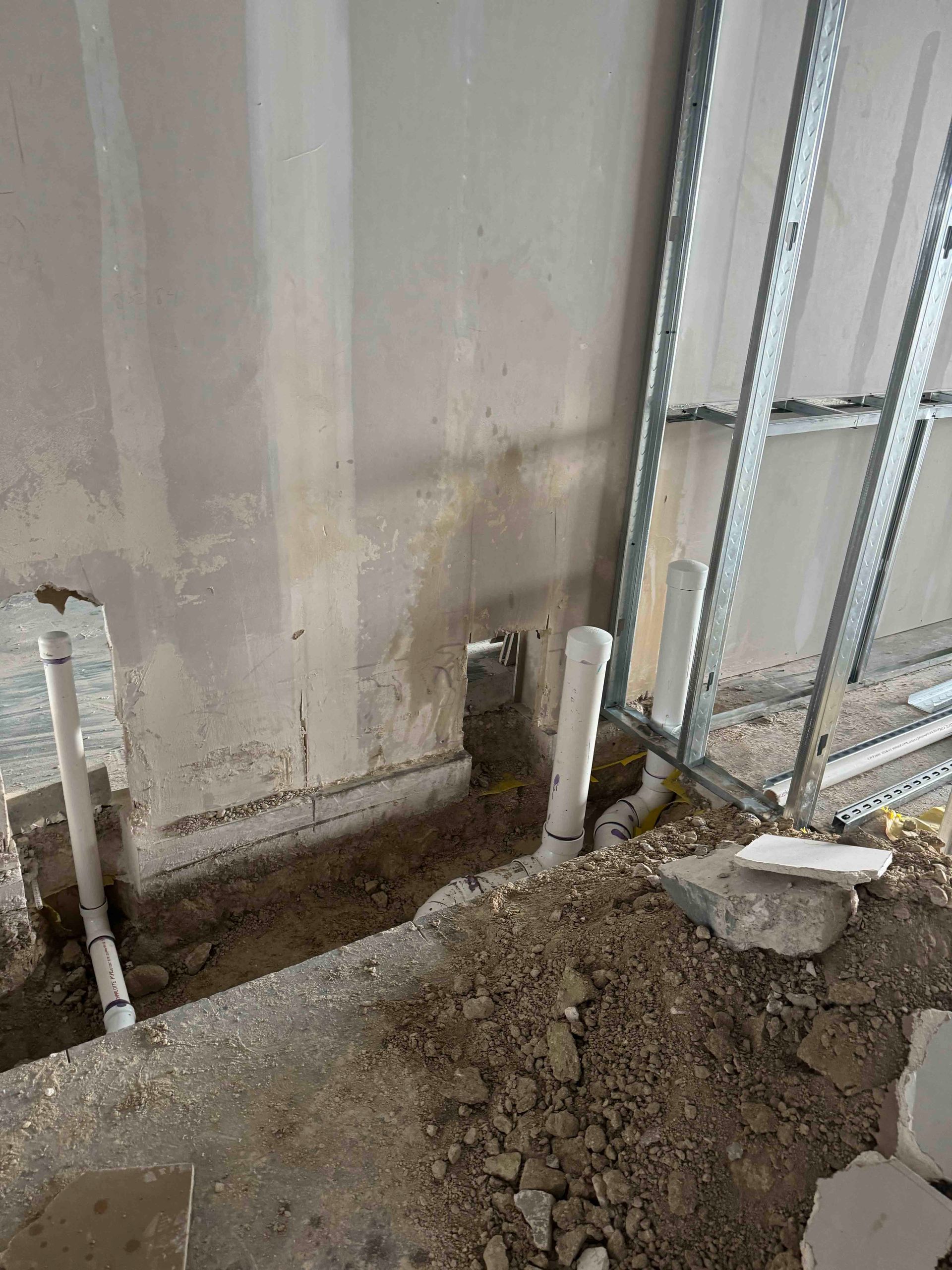 A hole in the ground next to a wall with pipes coming out of it.