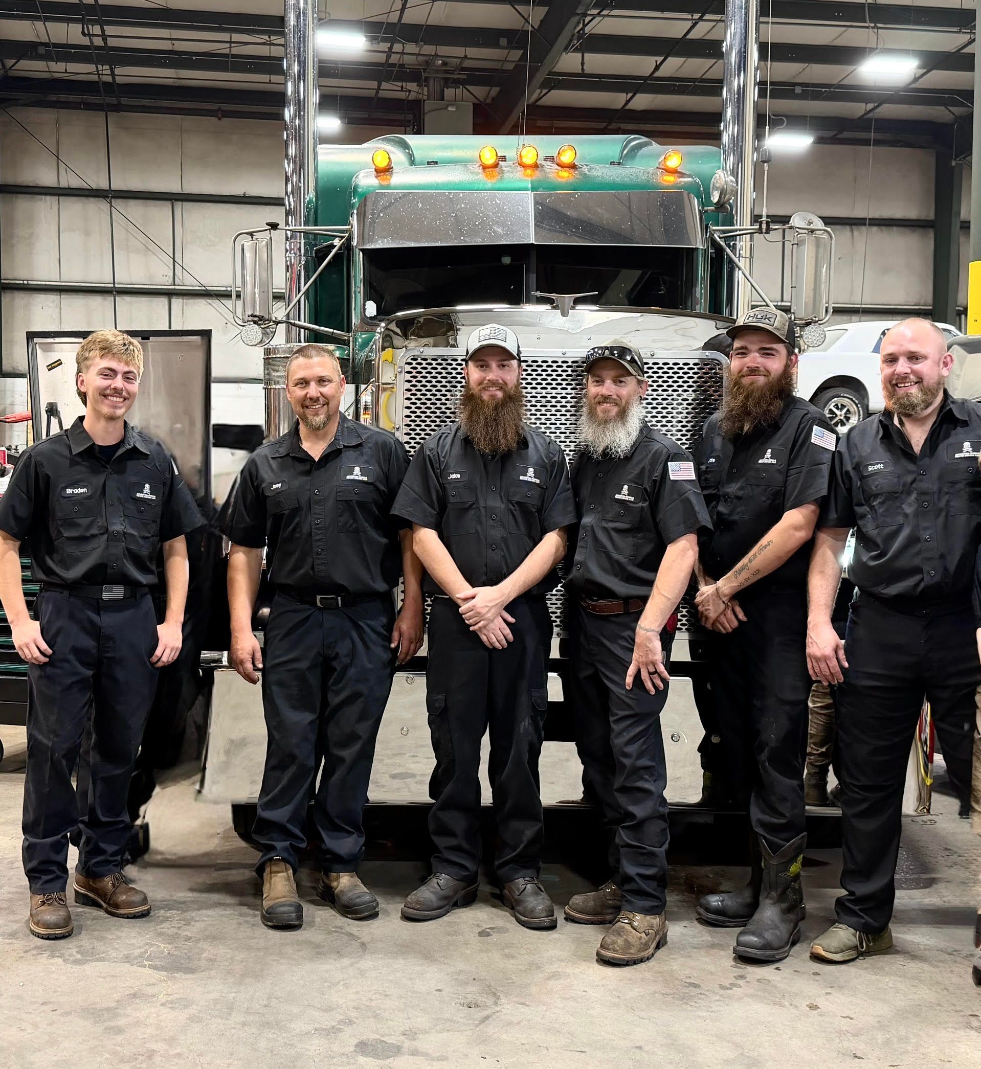 Six workers in black uniforms stand in a workshop in front of a green truck