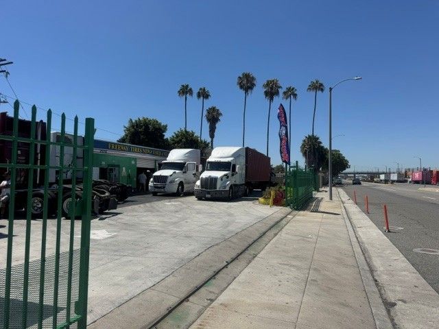 Trucks parked beside a fenced lot on a sunny roadside with palm trees and a clear blue sky.
