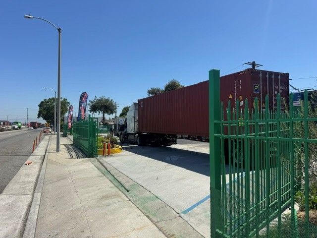 Long red freight truck parked beside a sidewalk and green fence on a sunny street