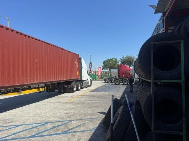 Shipping yard with red containers and trucks under a clear blue sky, with stacked pipes in the foreground