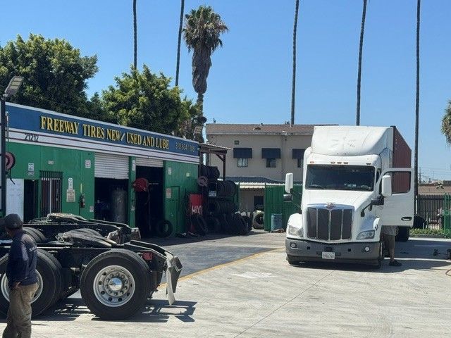 White semi-truck in a sunny lot beside a green storefront with palm trees and bare truck chassis in front