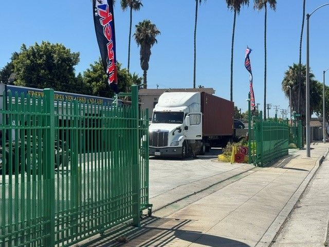 White semi-truck beside a green fence on a sunny sidewalk with palm trees and banners