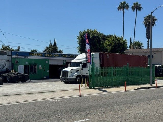 Green tire shop with two semi trucks, green fence, and tall palm trees under a clear blue sky.