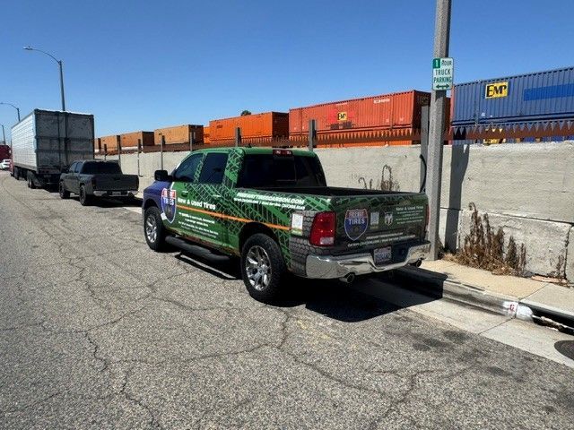 Green and black pickup truck parked on a street beside stacked shipping containers and a concrete wall.