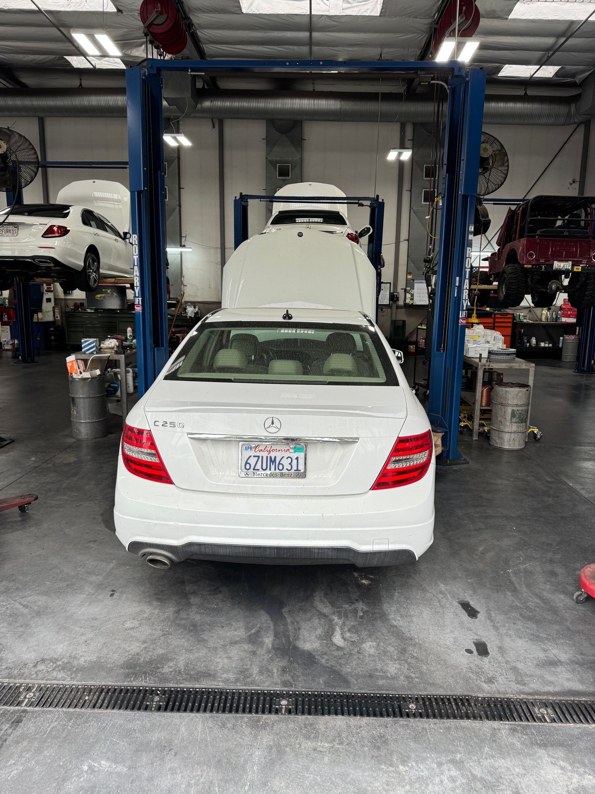White sedan on a lift in an auto repair shop, viewed from behind with hood and trunk open.