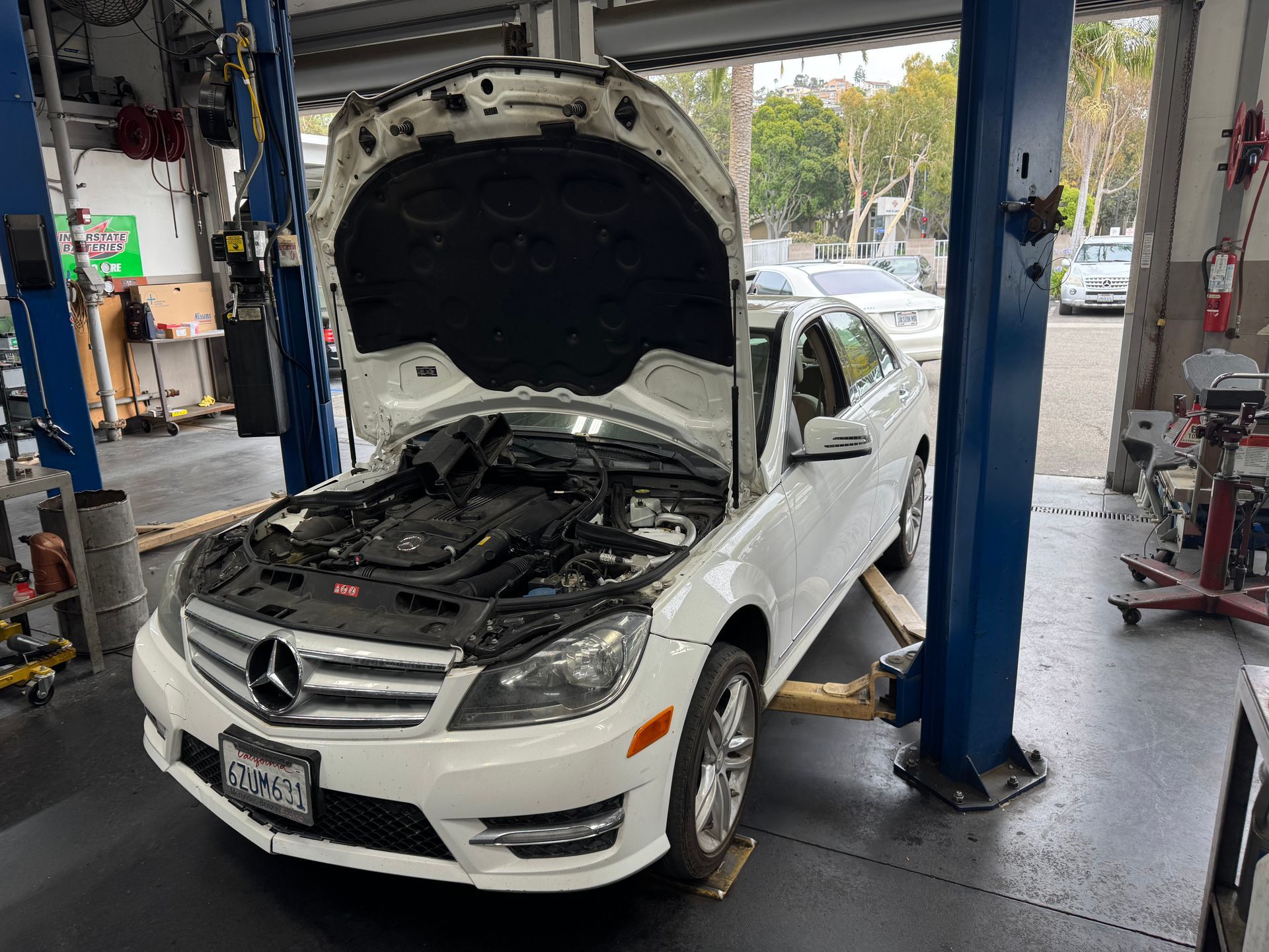 White Mercedes sedan with hood open in an auto repair shop, raised on a lift.