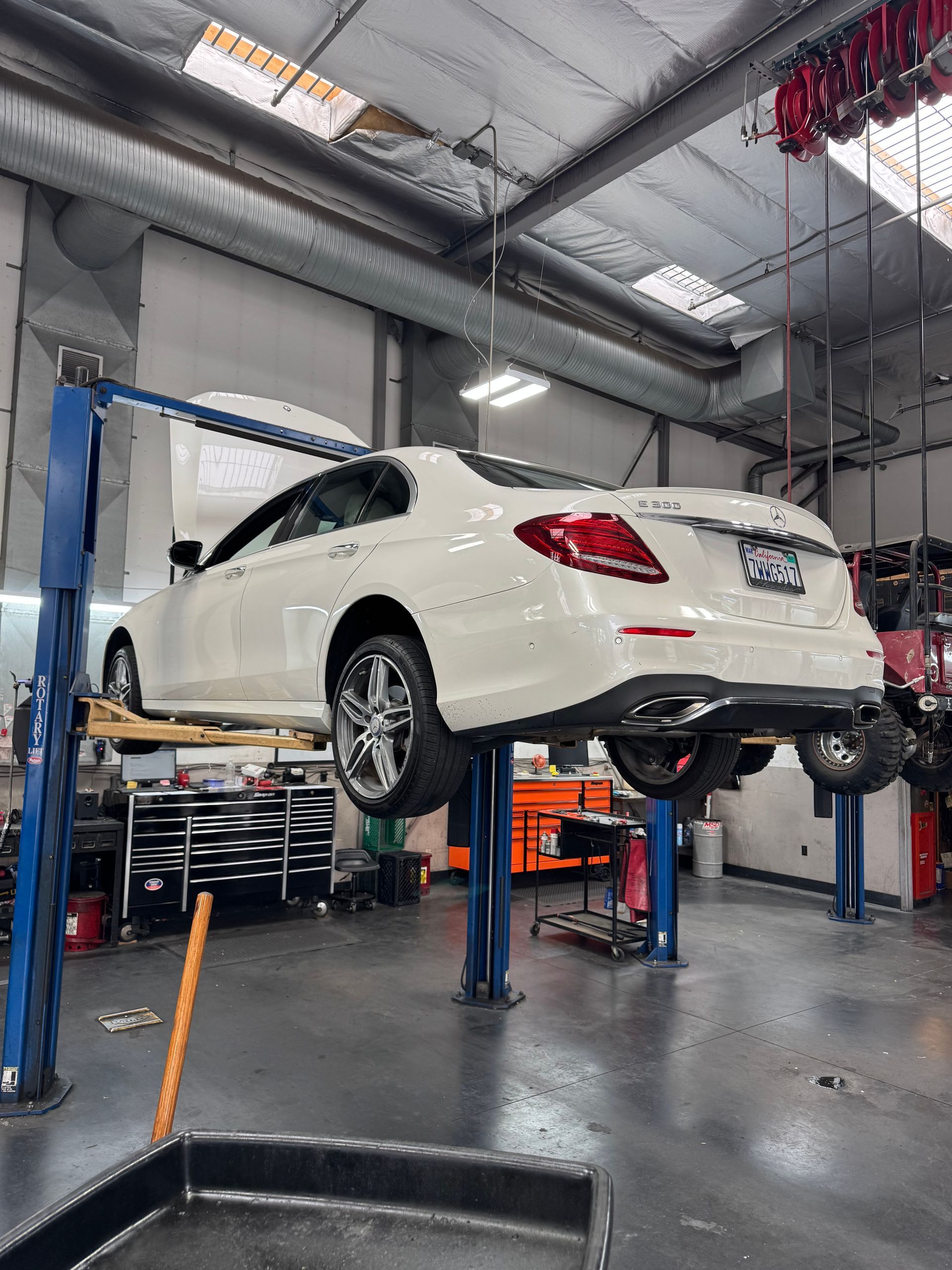 White sedan raised on a car lift inside an auto repair shop, viewed from the rear.