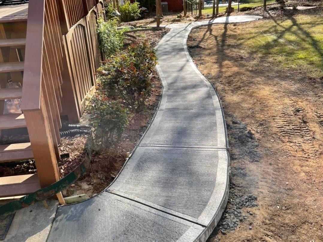 Curving concrete walkway in a yard, beside a wooden fence and shrubs, leading to a street.