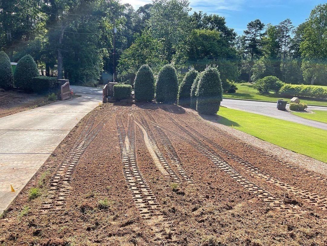 A freshly tilled brown garden bed with tire tracks leading to green shrubs and a driveway.
