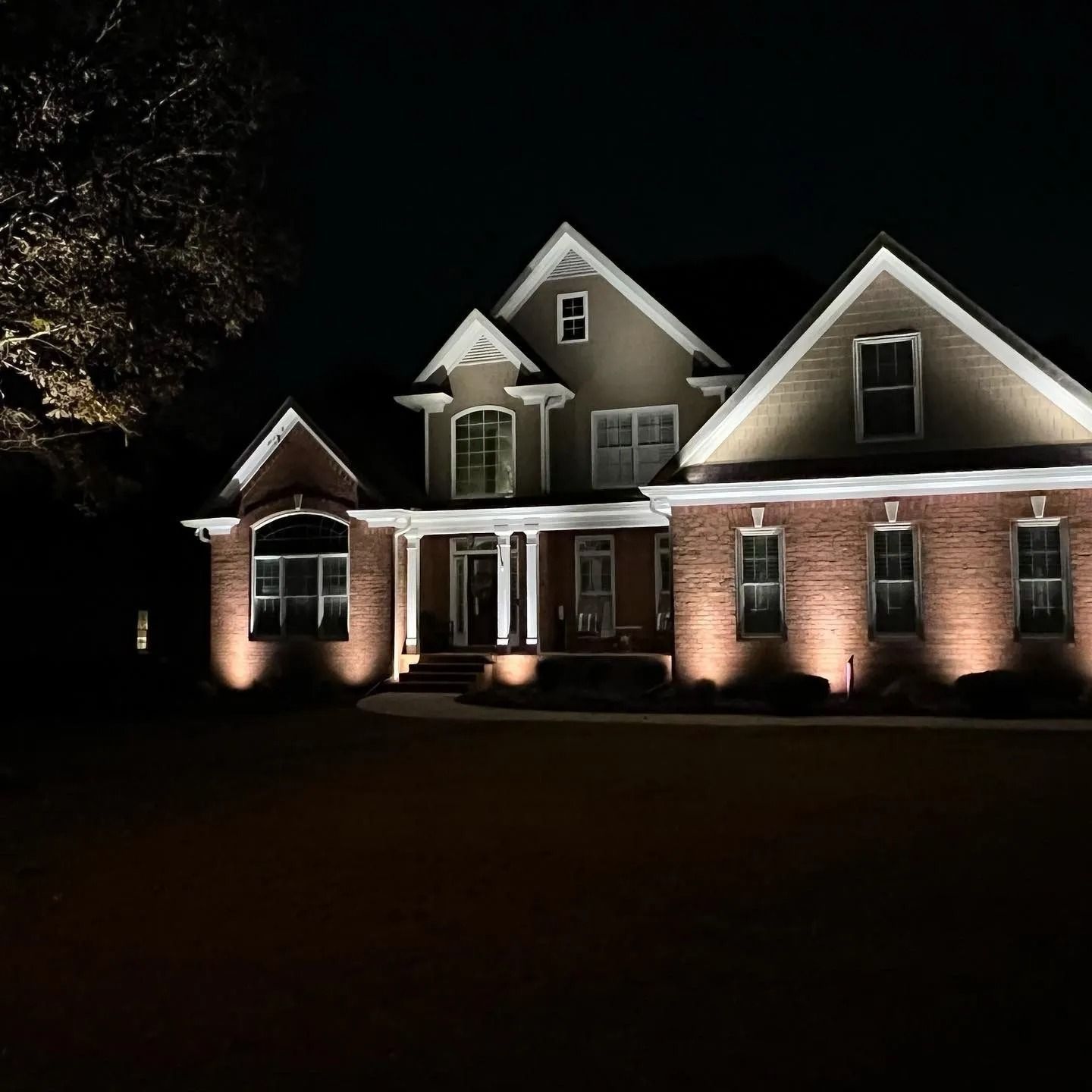 House exterior illuminated at night, featuring brick and tan siding, lit by spotlights.