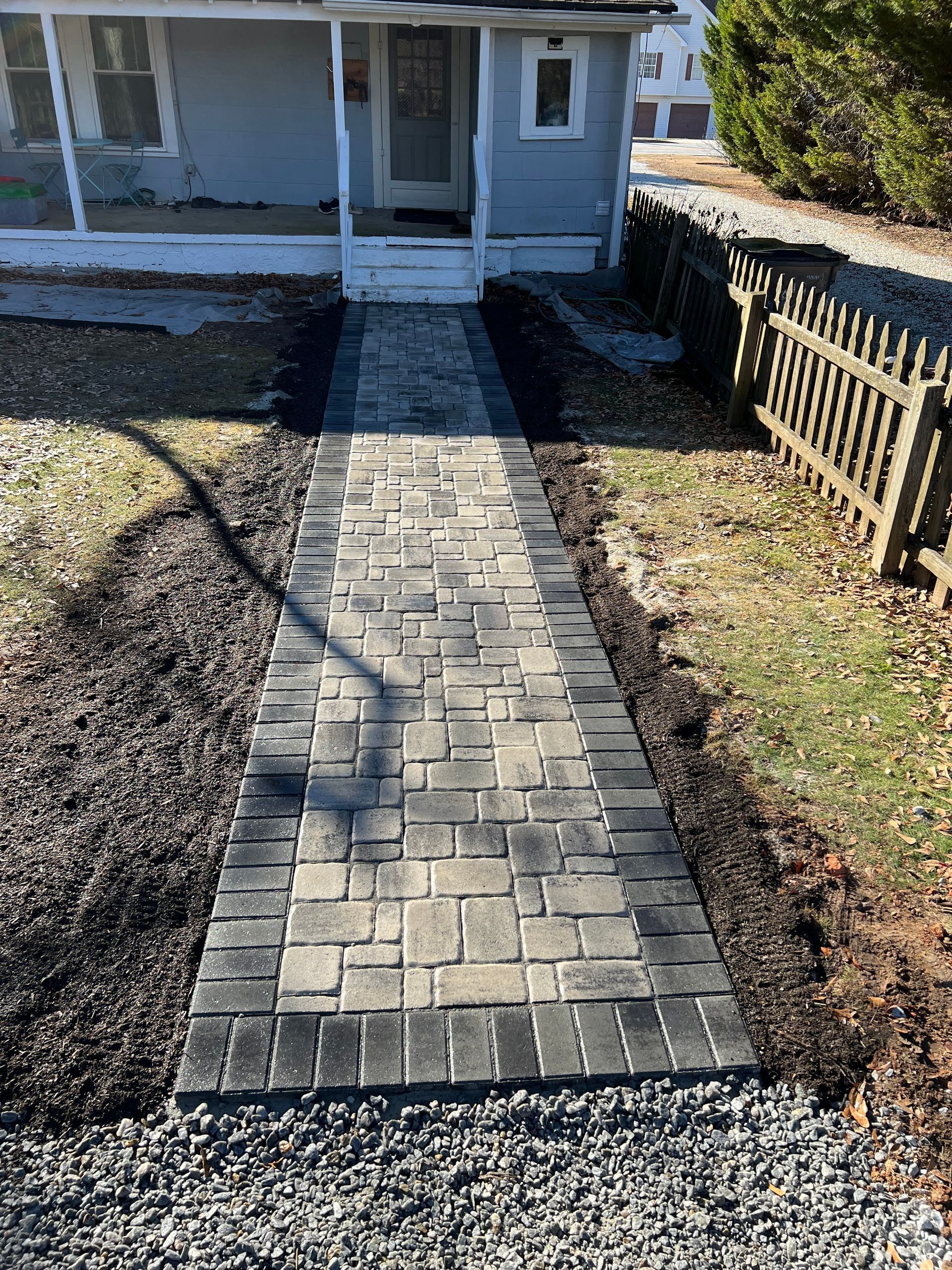 Brick walkway leading to a light blue house with a covered porch. Black mulch and a small fence border the path.