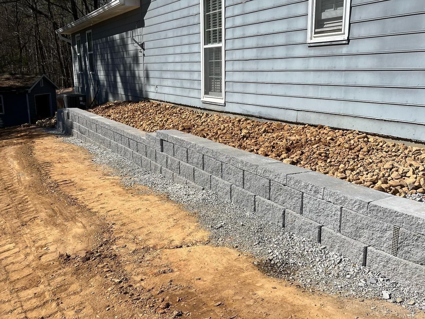 Gray retaining wall built in front of a blue house; brown gravel and dirt visible.