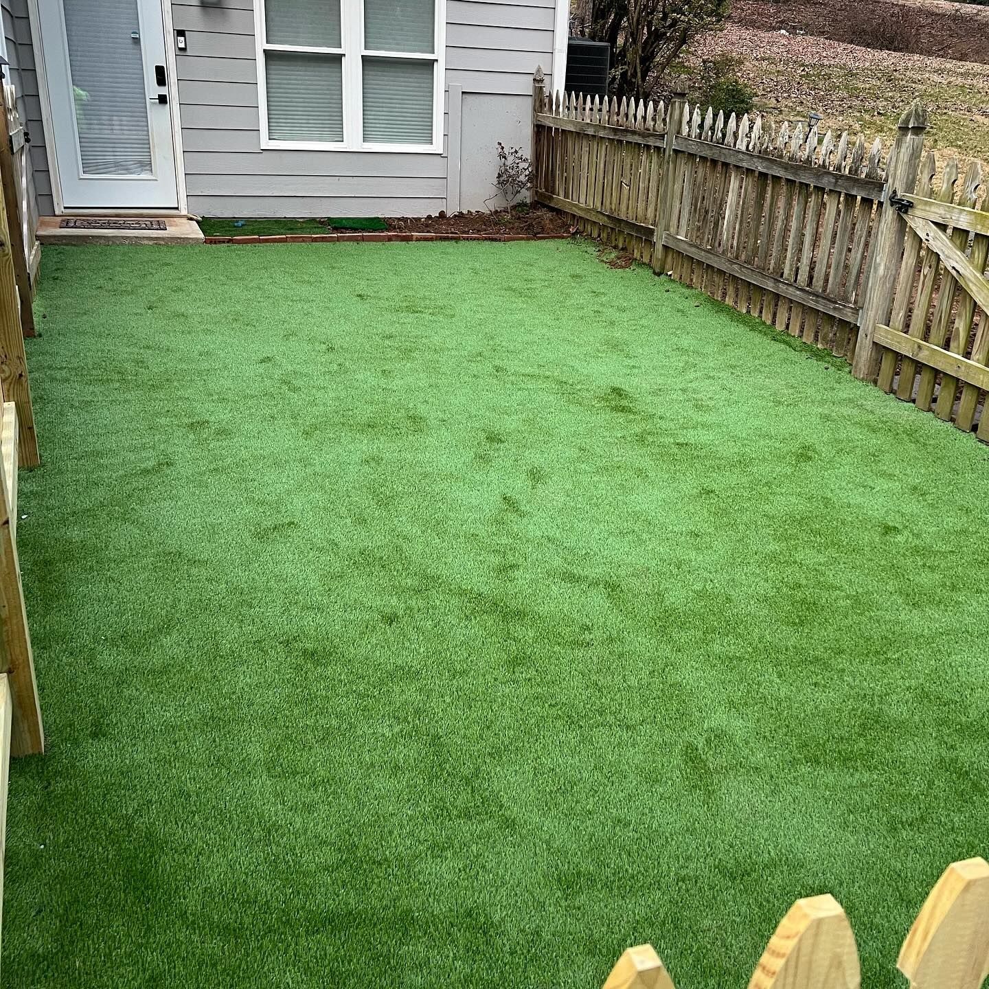 Green artificial turf in a small fenced yard with a door and a window in the background.