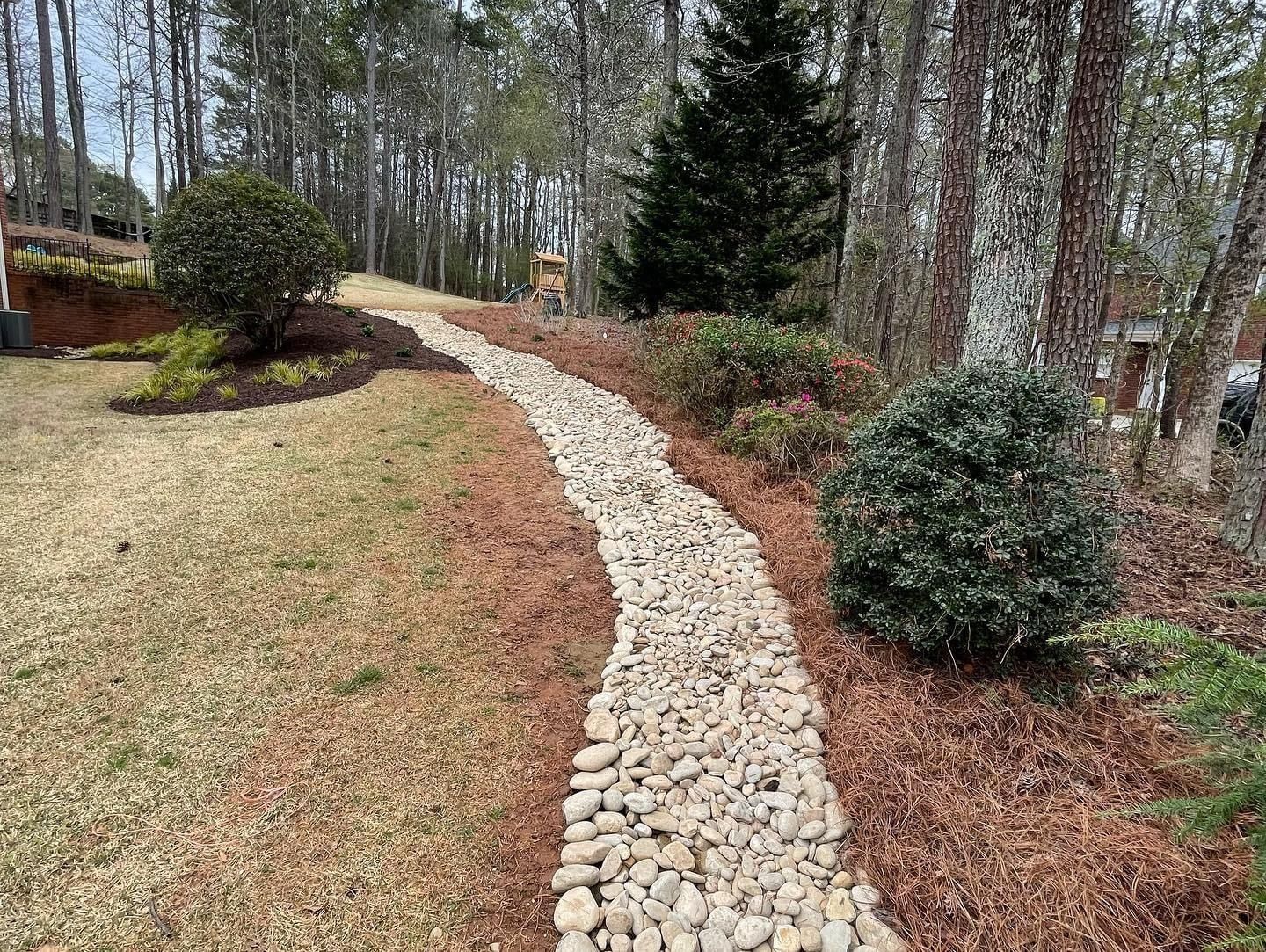 Gravel-lined drainage channel in a residential yard, bordered by brown pine straw and bushes, leading into a wooded area.