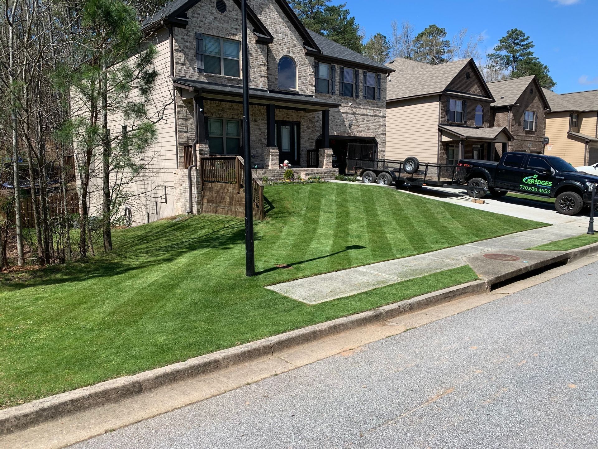 Lawn care scene: a two-story home with striped green lawn, black truck, and trailer parked next to a neighbor's house.