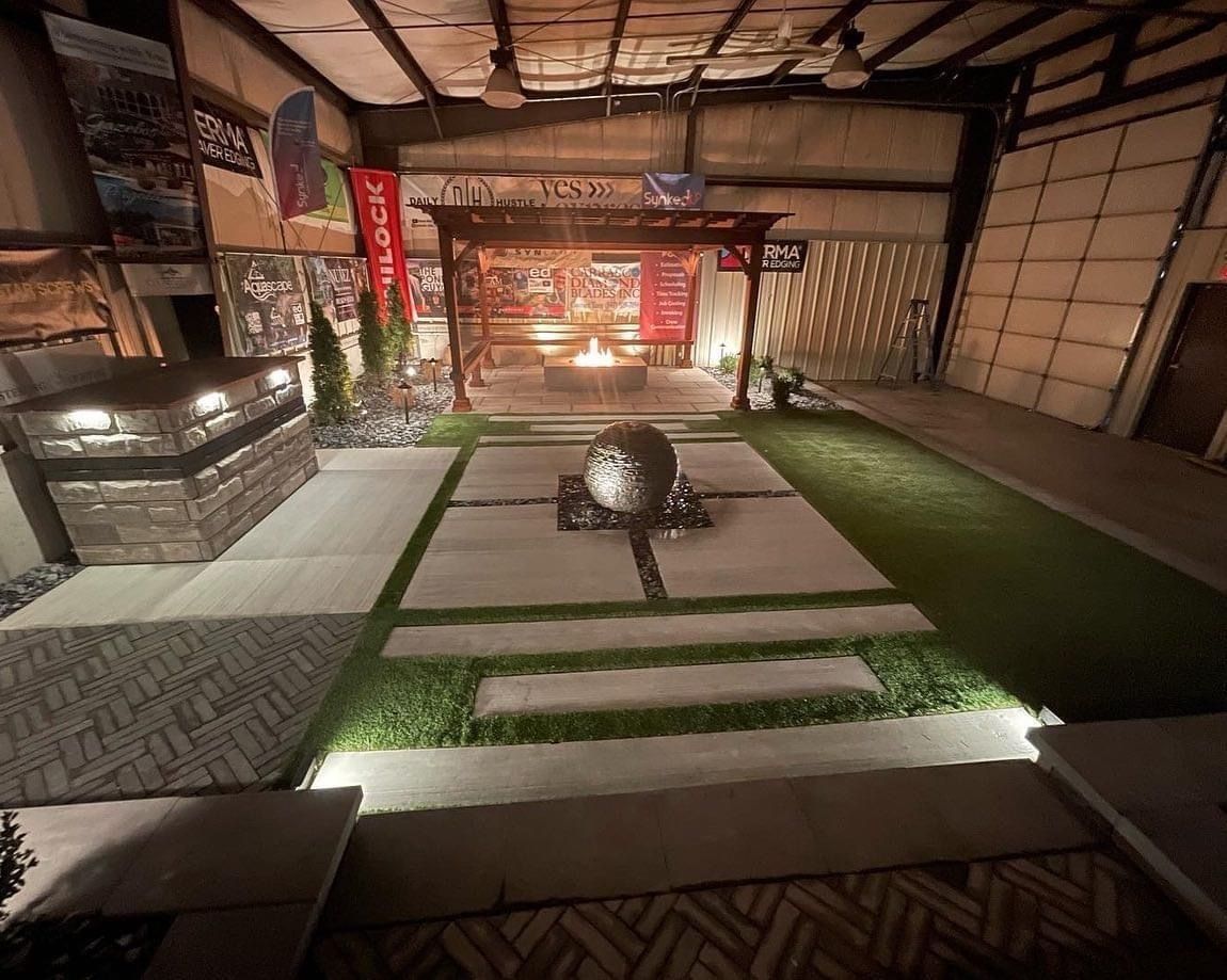 Display of outdoor landscaping elements, with pavers, greenery, and a fire pit under a pergola.