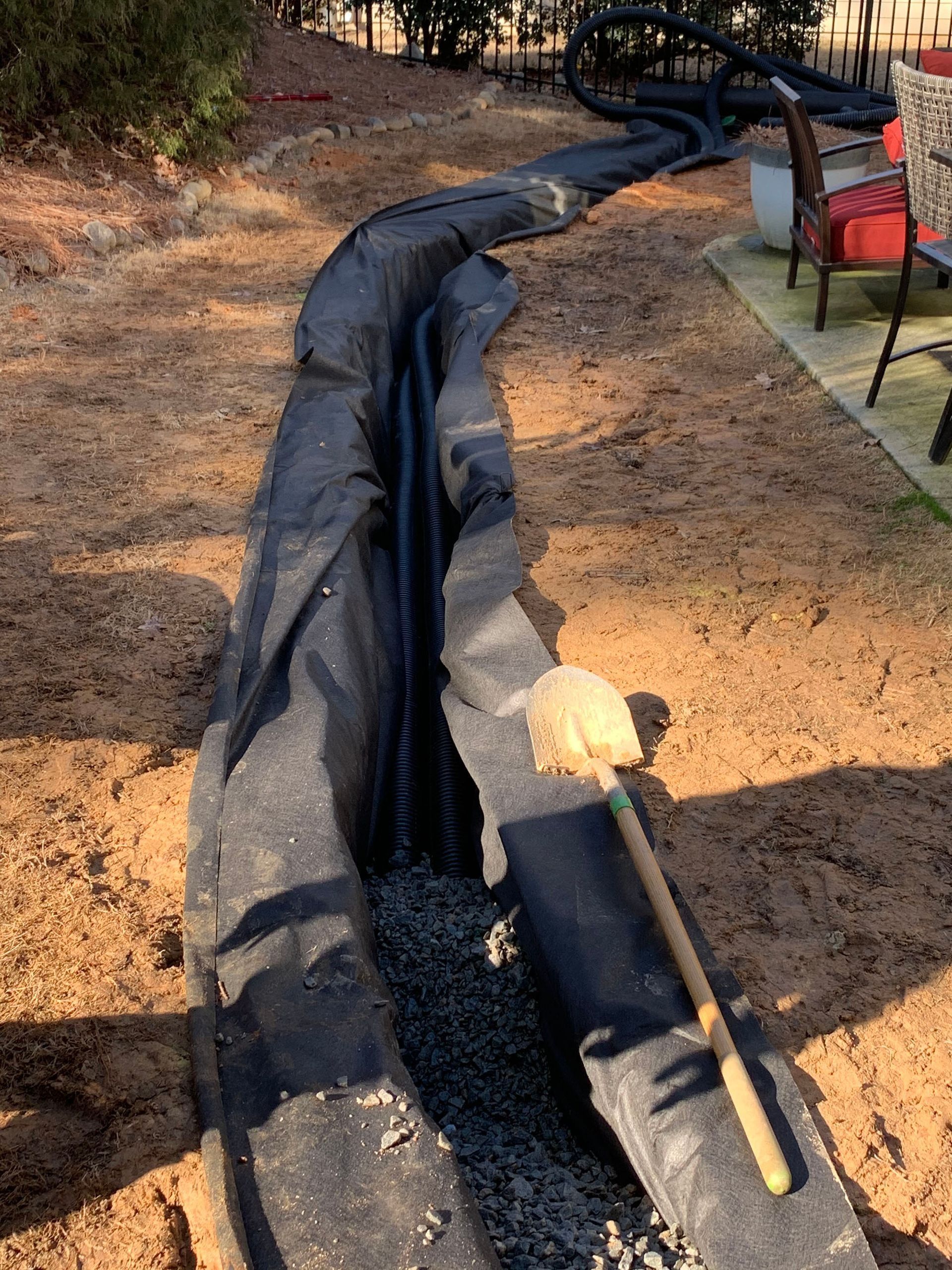 Trench with black fabric, gravel, and pipe, likely for drainage, runs along a sandy yard; shovel rests inside.