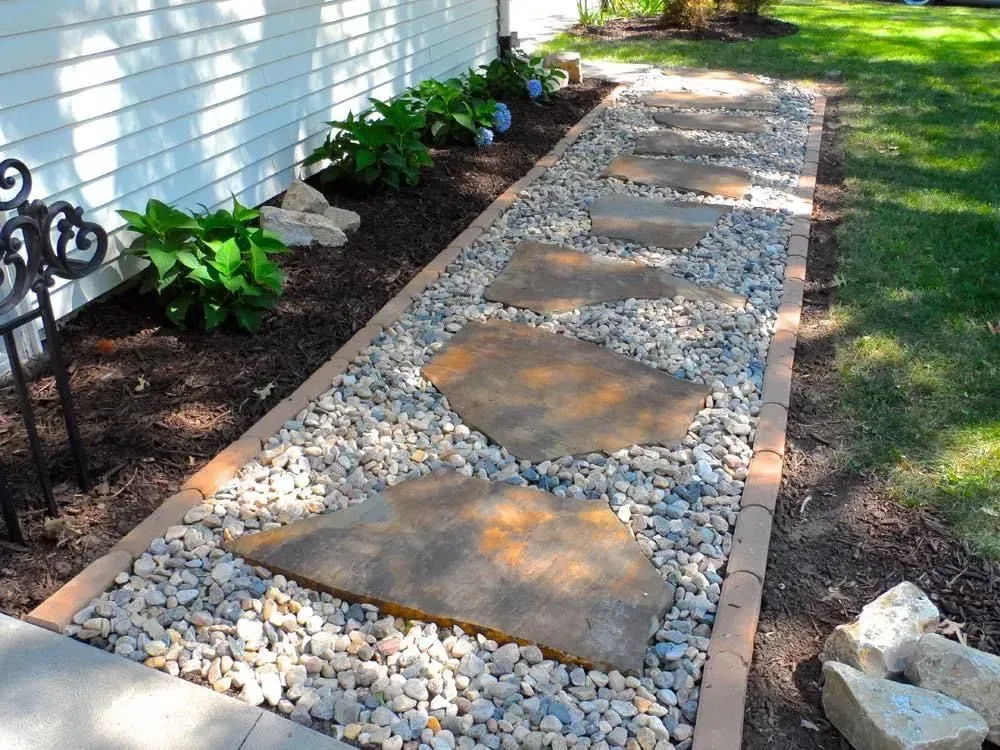 Stone pathway with stepping stones, bordered by rocks and brick, beside a house with green plants.
