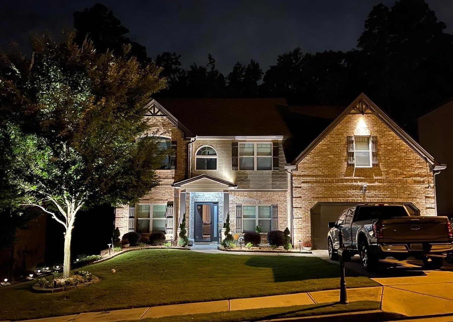 Two-story house illuminated at night with a truck in the driveway. Trees and lawn are also lit.