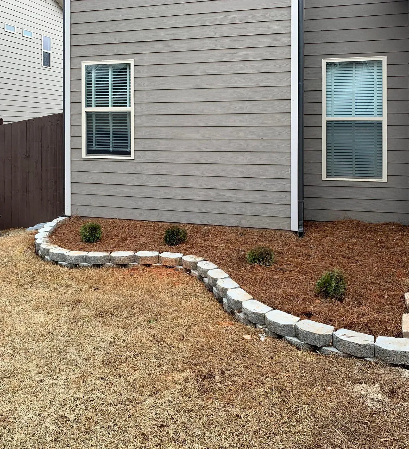 Exterior of a house with gray siding, two windows, and a curved brick border framing a flower bed with mulch.