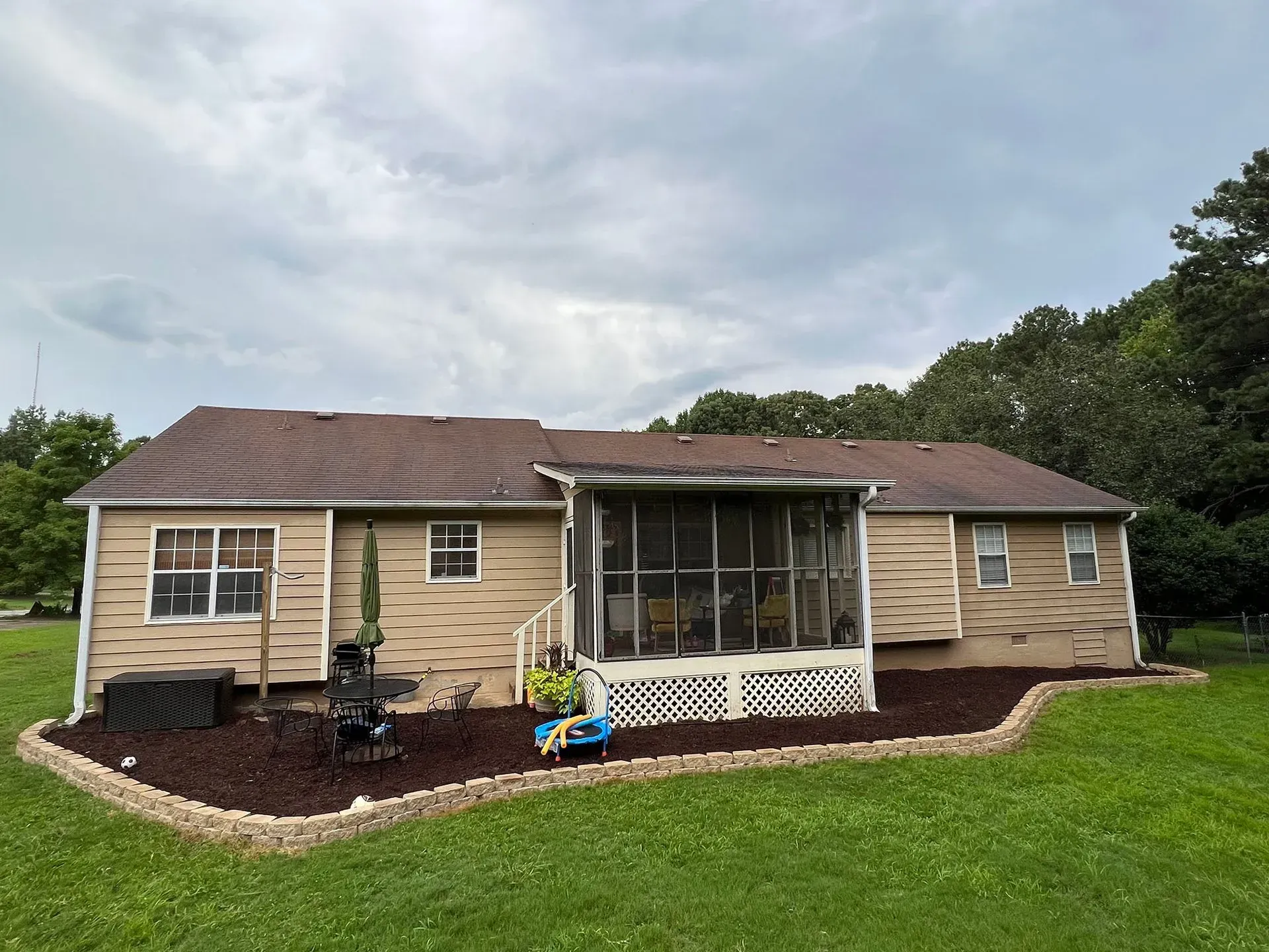 Back view of a tan house with a brown roof and a screened-in porch, surrounded by green grass and mulch landscaping.