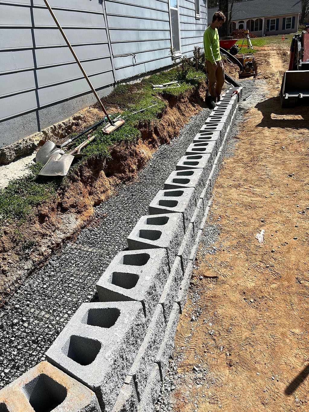 Construction of a retaining wall with cinder blocks beside a building; person operating a leaf blower.