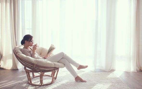 Woman reading a book and drinking tea in a sunny room, seated in a wicker chair near a window.