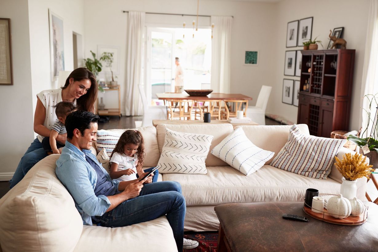 Family of four relaxing in a bright living room, man and child on sofa with tablet, woman holding toddler.