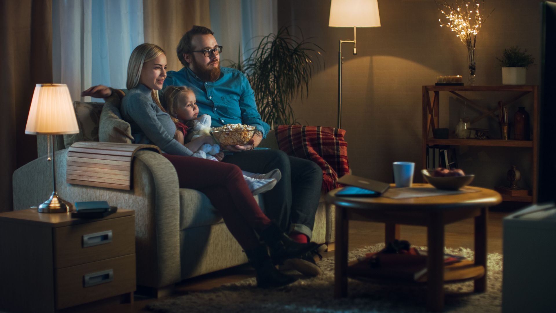 Family of three watching TV, sharing popcorn on a couch in a cozy, dimly lit living room.